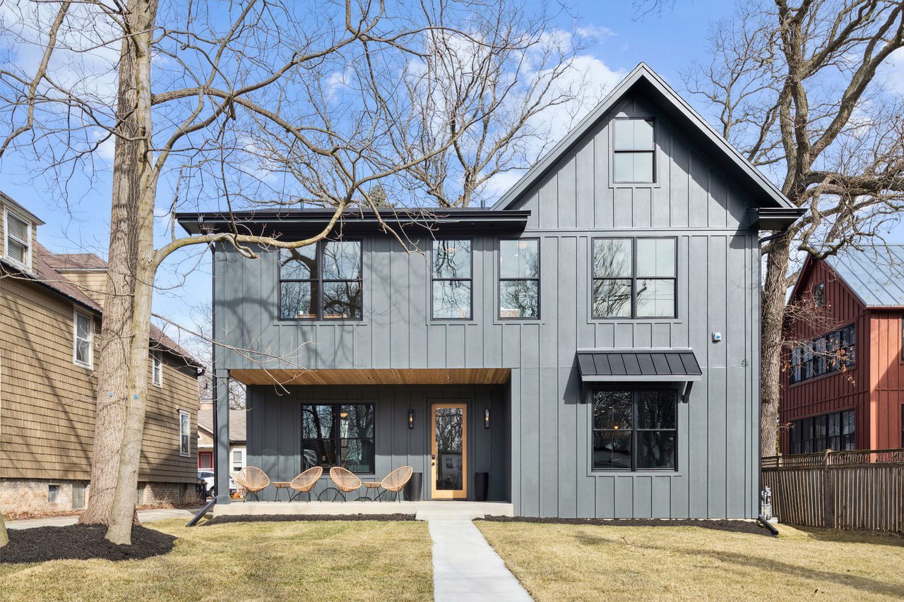 20140825_100318 Front view of a modern two-story house with gray vertical siding, multiple windows, and a covered porch with seating. A walkway leads to the front door, and there are trees and neighboring homes in the background.